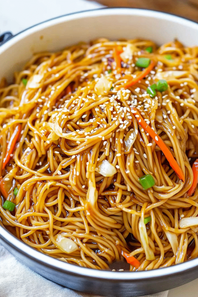 Bowl of glossy teriyaki noodles with vegetables and sesame seeds, garnished with green onions and chopsticks on the side